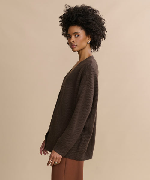 A woman with short curly hair stands in profile before a beige background, wearing the Cashmere Cocoon Cardigan and brown pants. She gazes calmly at the camera with a neutral expression.