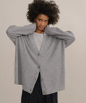 A person with curly hair wears the loose, gray Cashmere Cocoon Cardigan over a light shirt and black pants, standing with both hands touching the back of their neck against a plain background.