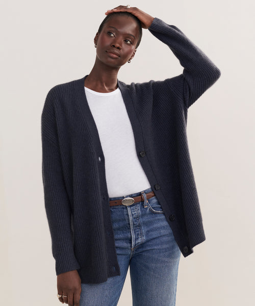 A person wears the Cashmere Cocoon Cardigan in dark blue over a white t-shirt and blue jeans, standing with one hand on their head against a plain background.