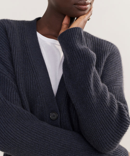 A person wearing the Cashmere Cocoon Cardigan in dark blue over a white shirt rests their chin on their hand, partially covering their face. The cardigan features a ribbed texture and visible button detail.