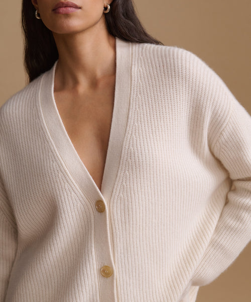 A close-up of a woman wearing a cream-colored ribbed knit cardigan with a V-neck and buttons, set against a warm beige background.