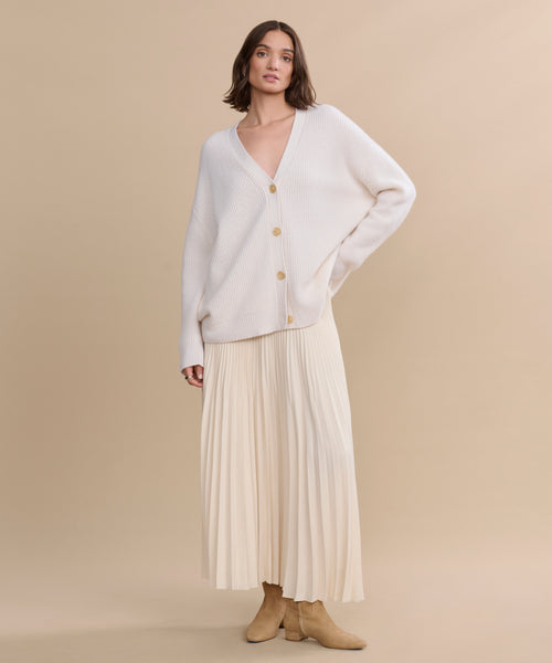 A person stands against a beige background wearing the oversized, lightweight Cashmere Cocoon Cardigan with large buttons, a matching pleated midi skirt, and beige ankle boots.