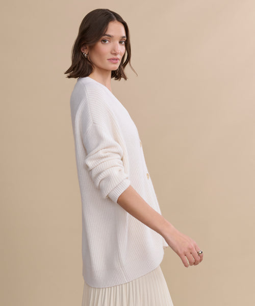A woman with shoulder-length brown hair stands in profile against a beige background, wearing an oversized Cashmere Cocoon Cardigan in cream and a light pleated skirt.