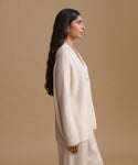 A woman in side profile wearing a cream ribbed knit cardigan and matching cream trousers, set against a solid beige studio background.