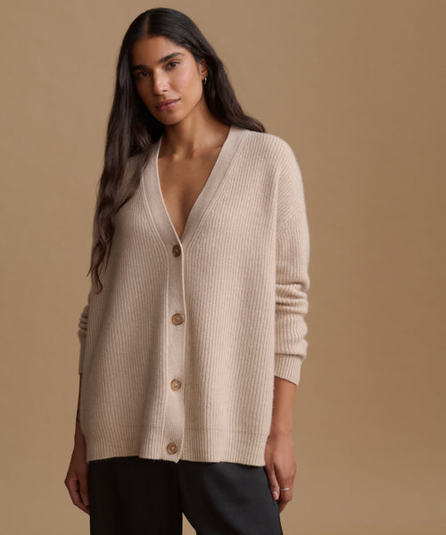 A woman modeling a beige ribbed knit cardigan with brown buttons, paired with black pants, against a solid warm beige studio background.