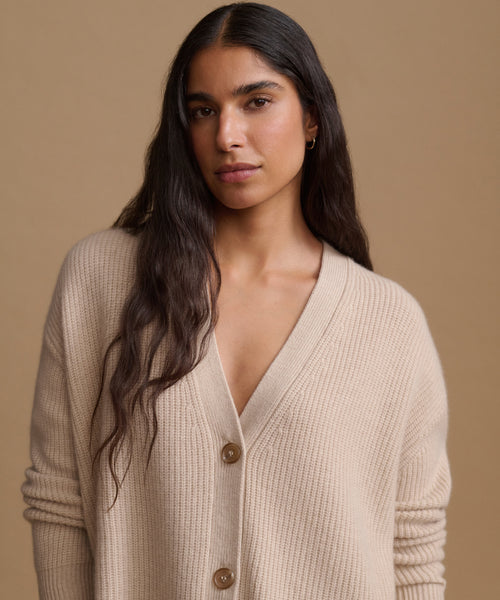 A woman with long dark hair wearing a beige cardigan with buttons, set against a solid warm beige background.