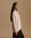 A woman in side profile wearing a ribbed cream-colored cardigan with buttons and black pants, set against a solid beige studio background.