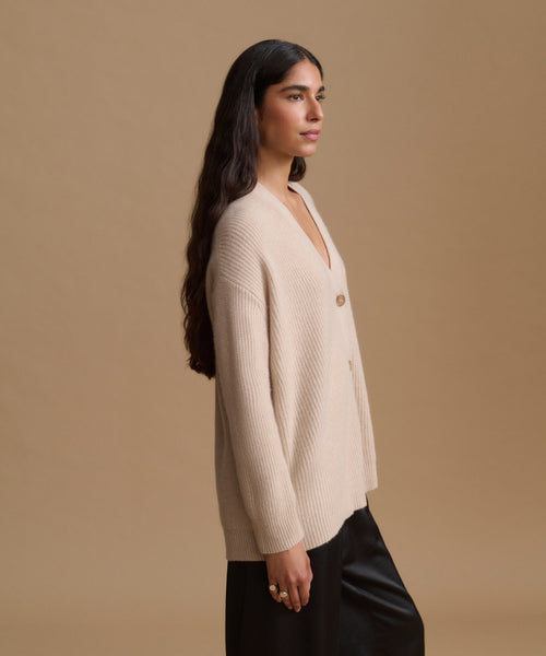 A woman in side profile wearing a ribbed cream-colored cardigan with buttons and black pants, set against a solid beige studio background.