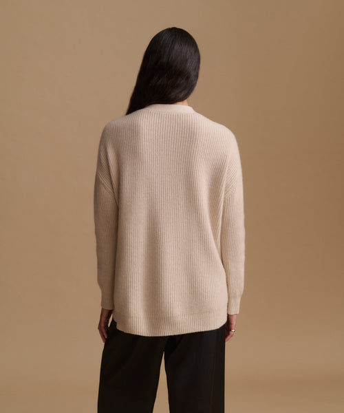A woman viewed from behind wearing a ribbed knit sweater in a light cream color and black wide-leg pants, set against a solid warm beige studio background.