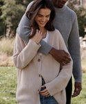 A woman wearing a cream ribbed knit button-front cardigan over a white top, with a man in a gray ribbed sweater standing behind her in an outdoor setting.