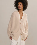 A woman models the Cashmere Cocoon Cardigan in beige over a cream blouse and white pants, standing against a neutral background with her hands in her pockets, gazing to the side.