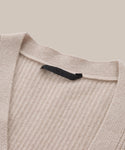 Close-up of the Cashmere Cocoon Cardigan, featuring beige ribbed knit fabric, a V-neckline, and a black label at the back of the neck. The background is solid light beige.