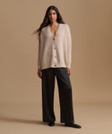 A woman stands against a warm beige studio background wearing a light cream knit cardigan with brown buttons and black wide-leg trousers.