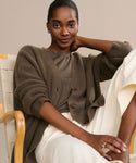 A woman with dark skin and pulled-back hair sits in a wooden chair, smiling softly at the camera while wearing the Cashmere Cocoon Cardigan over a brown top and white pants, her elbow resting on the chair.
