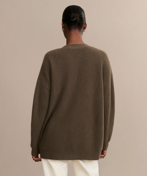 Seen from behind, a person with dark hair in a low bun wears the Cashmere Cocoon Cardigan in ribbed brown with white pants, standing against a plain beige background.