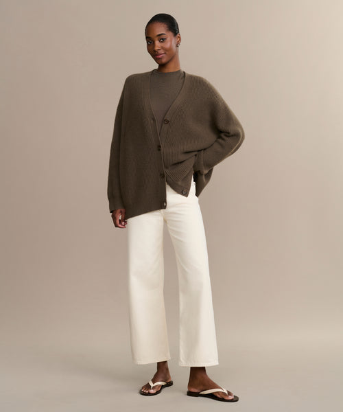 A person stands against a beige background wearing the Cashmere Cocoon Cardigan, a matching brown top, white wide-leg pants, and brown sandals. Their short hair is pulled back, and they are smiling slightly.