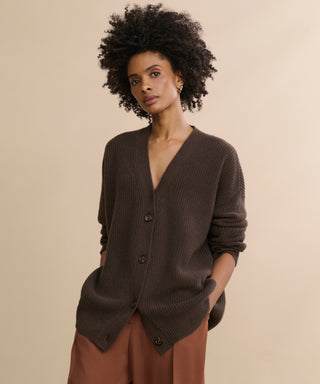 A woman with curly dark hair wears the oversized Cashmere Cocoon Cardigan in dark brown and rust-colored pants, standing against a plain beige background with her hands in her pockets.