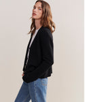 A person with long brown hair models the relaxed fit Cashmere Colette Cardigan in black, layered over a white top and blue jeans, standing sideways and facing the camera against a plain light background.