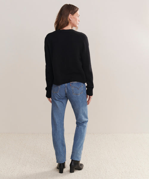 A person with light brown hair stands facing away from the camera against a neutral background, wearing the black Cashmere Colette Cardigan in a relaxed fit, paired with blue jeans and black ankle boots.