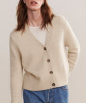 A person wearing a relaxed fit Cashmere Colette Cardigan in oatmeal over a white shirt and blue jeans, standing against a plain, light background.