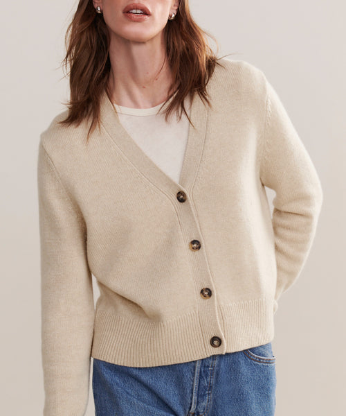 A person wearing a relaxed fit Cashmere Colette Cardigan in oatmeal over a white shirt and blue jeans, standing against a plain, light background.