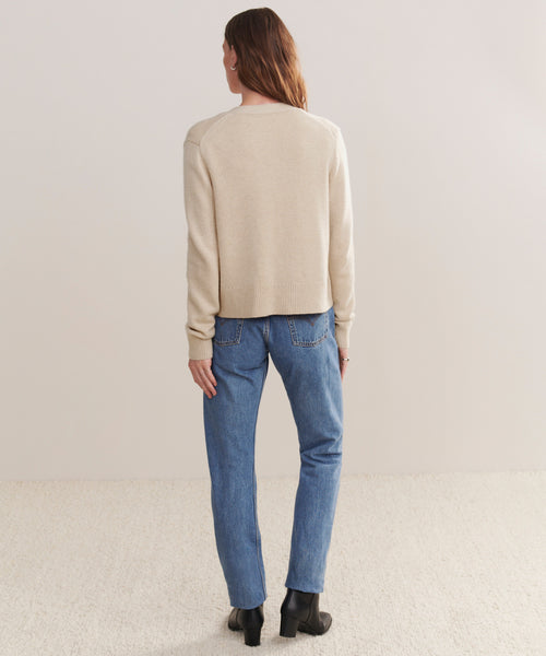 A person with long brown hair stands facing away, wearing the Cashmere Colette Cardigan, blue jeans, and black heeled boots against a plain light background and textured carpet.