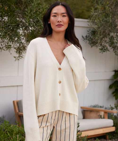 A woman with long dark hair wears the Cashmere Colette Cardigan in ivory and beige pants with black vertical stripes, standing outdoors by a white fence and green foliage, with a wooden bench in the background.