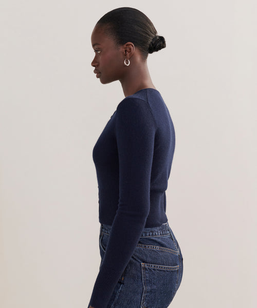 A woman with her hair in a neat bun is shown in profile, wearing the Cashmere Finley Cardigan in dark navy with high-waisted blue jeans, standing against a plain light background.