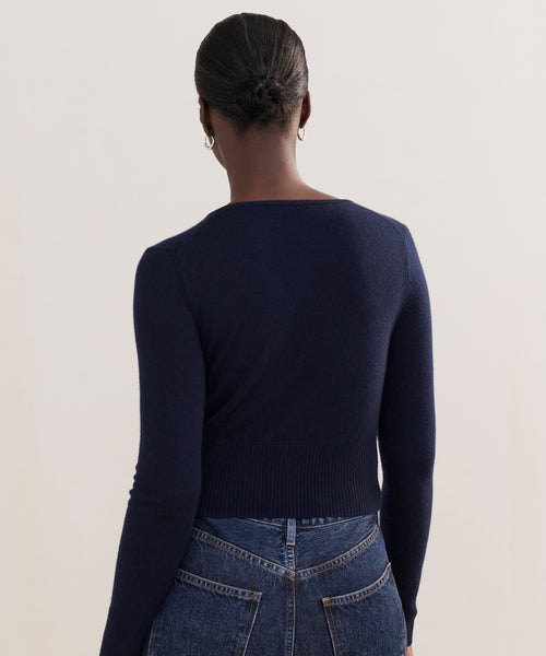 A person with their hair in a neat bun is seen from behind, wearing a dark navy Cashmere Finley Cardigan paired with high-waisted blue jeans, set against a plain light background.