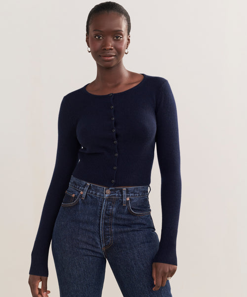 A woman wears the shrunken fit Cashmere Finley Cardigan in dark navy and high-waisted dark blue jeans, standing against a plain light background and looking confidently at the camera.