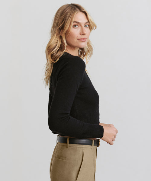 A woman with wavy blonde hair wears the Cashmere Finley Cardigan in black and beige pants, standing in profile against a plain light background and looking toward the camera.