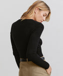 A woman with shoulder-length blonde hair wears the shrunken fit black Cashmere Finley Cardigan with button front detailing, paired with tan pants and a black belt, looking down over her shoulder against a plain light background.