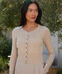 A woman with long dark hair wears the Cashmere Finley Cardigan with fine buttons and a skirt, standing outdoors in sunlight and greenery, looking slightly right with a calm expression.