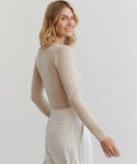 A woman with shoulder-length blonde hair smiles over her shoulder, wearing the Cashmere Finley Cardigan in beige with fine button detailing and high-waisted light pants against a plain light background.