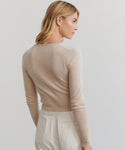A blonde woman stands in profile, facing left, wearing the beige Cashmere Finley Cardigan with fine buttons and light pants against a plain, light background.