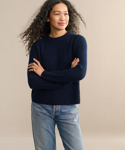 A woman with long curly hair stands with arms crossed, smiling slightly against a plain beige background, wearing a classic dark blue Cashmere Fisherman Sweater and light blue jeans.