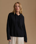 A woman wearing a black ribbed knit hooded sweater with drawstrings and light beige wide-leg trousers, standing against a solid warm beige studio background.