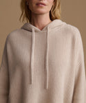A close-up of a woman wearing a ribbed knit hooded sweater in a light beige color, set against a solid warm tan background.