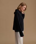 A woman wearing a black ribbed knit hooded cardigan and white wide-leg trousers, standing against a solid beige studio background.