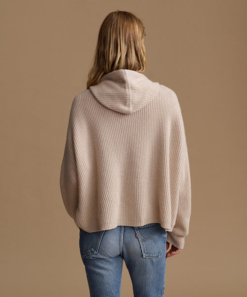 A woman viewed from behind wearing a beige ribbed knit hooded sweater and blue jeans against a solid tan background.