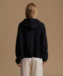 A woman viewed from the back wearing a black ribbed knit hooded sweater and cream wide-leg trousers, set against a solid warm tan studio background.
