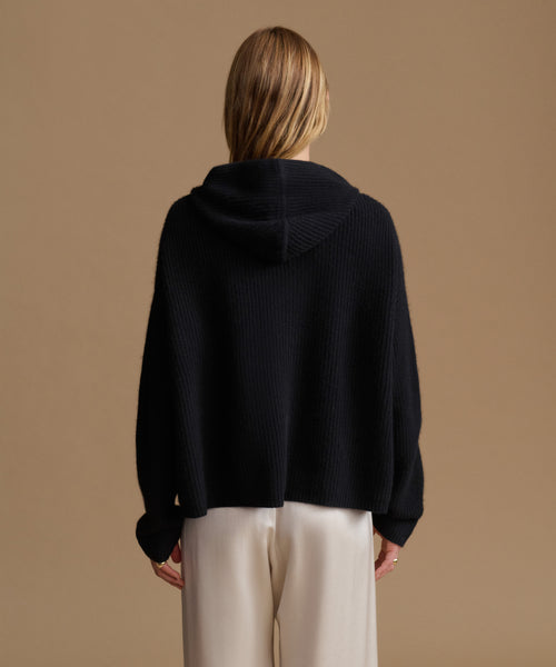 A woman viewed from the back wearing a black ribbed knit hooded sweater and cream wide-leg trousers, set against a solid warm tan studio background.