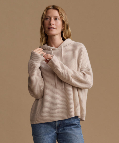A woman wearing a beige ribbed knit hoodie sweater and blue jeans, posing against a solid warm beige background.