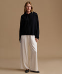 A woman stands against a warm beige studio background wearing a black oversized pullover hoodie, wide-leg cream pants, and black flats.