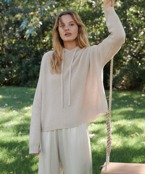 A woman stands outdoors on a grassy lawn holding a rope swing, wearing a cream-colored ribbed knit hoodie and matching cream pants.