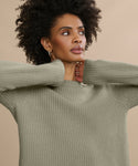 A woman with curly hair wears the Cashmere Fisherman Sweater in light olive green, posing with her hands near her neck against a beige background.