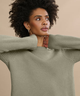 A woman with curly hair wears the Cashmere Fisherman Sweater in light olive green, posing with her hands near her neck against a beige background.