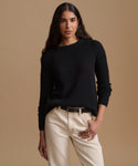 A woman modeling a black ribbed knit crewneck sweater, paired with cream-colored pants and a black belt, against a solid beige studio background.