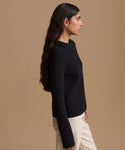 A woman in profile wearing a black ribbed knit sweater and cream trousers, standing against a solid tan studio background.