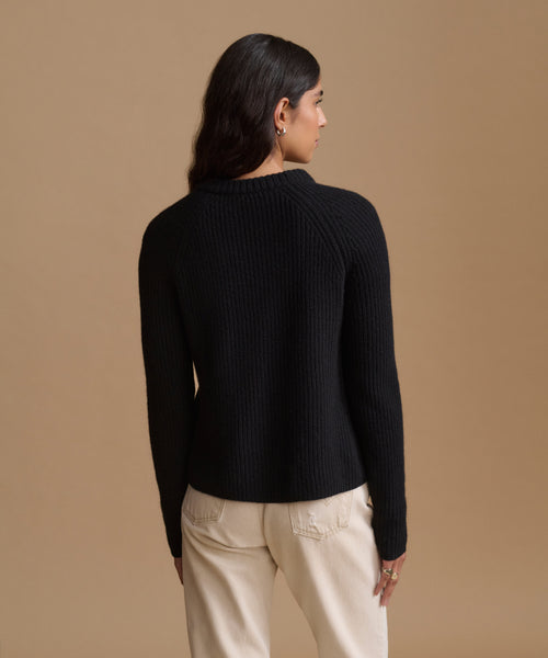 A woman shown from the back wearing a black ribbed knit sweater and cream pants, standing against a solid beige studio background.